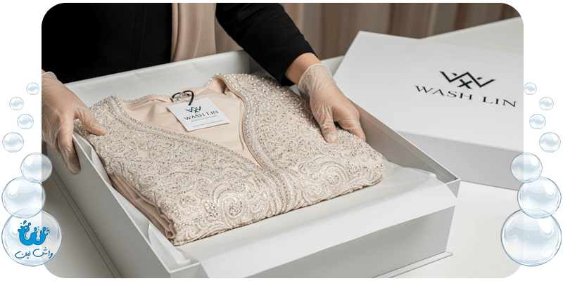 متخصص واش‌لین (خانم با حجاب) در حال بسته‌بندی لباس عروس تمیز شده در جعبه مخصوص بایگانی.